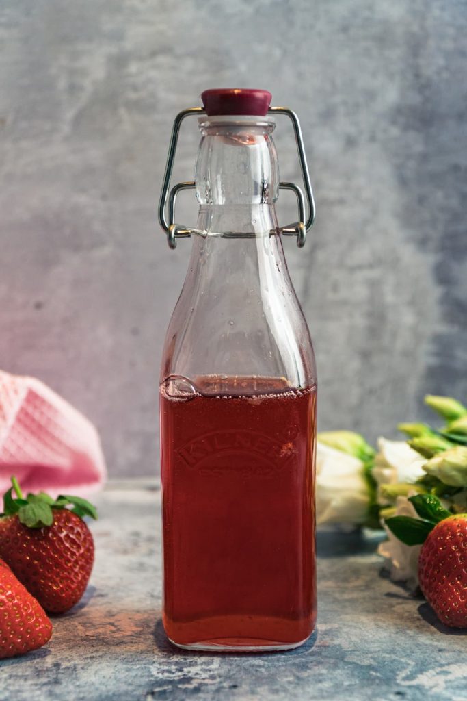 Bottle of strawberry syrup with strawberries, flowers and pink tea towel.